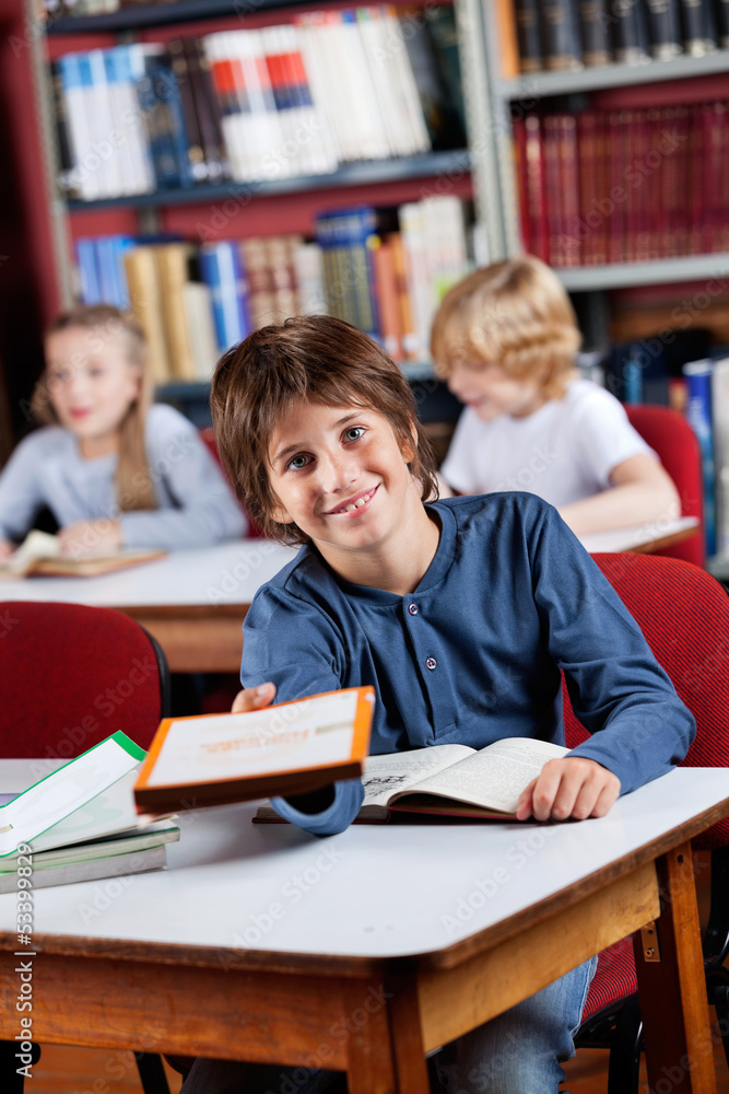 Happy Schoolboy Giving Book While Sitting At Table In Library Happy Schoolboy Giving Book While Sitting At Table In Library
