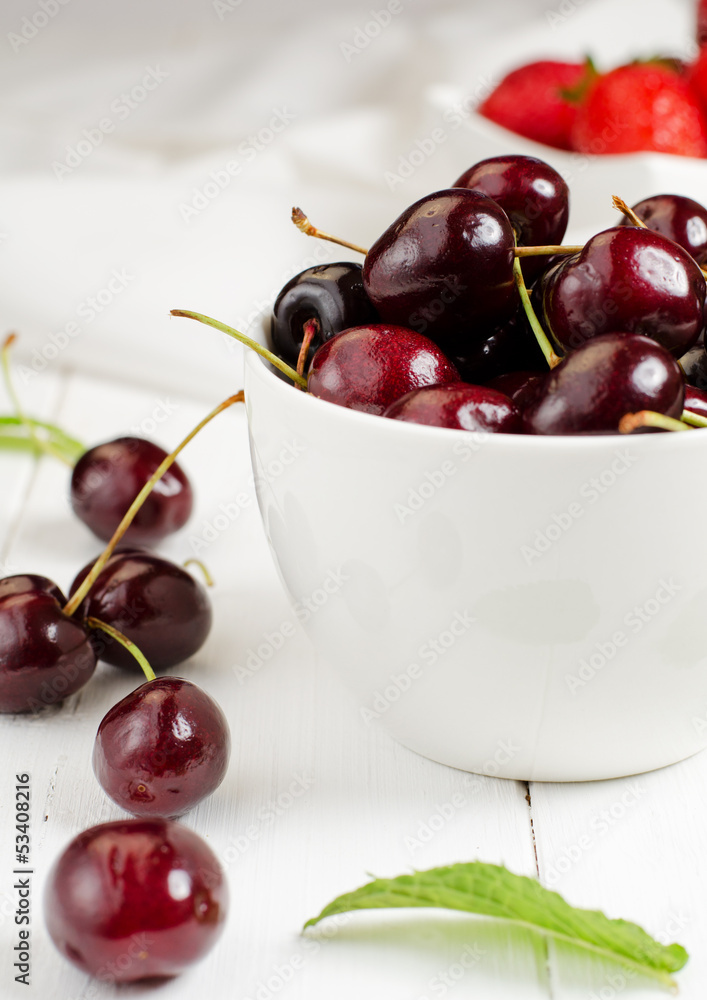 Cherries in the white bowl with strawberries in the background Cherries in the white bowl with strawberries in the background