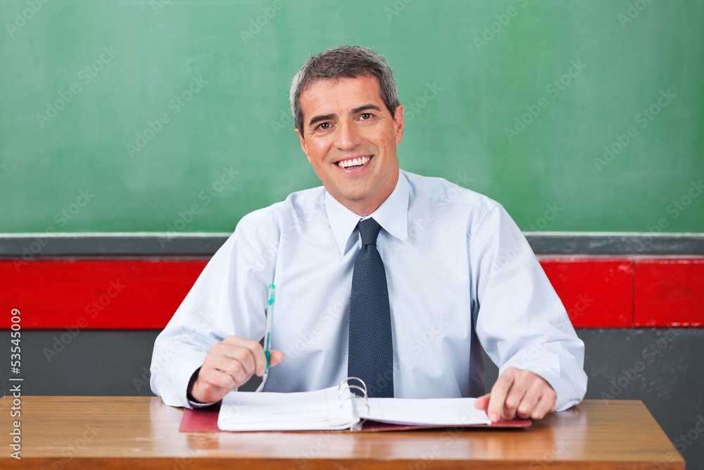 Happy Male Teacher With Pen And Binder Sitting At Desk Happy Male Teacher With Pen And Binder Sitting At Desk