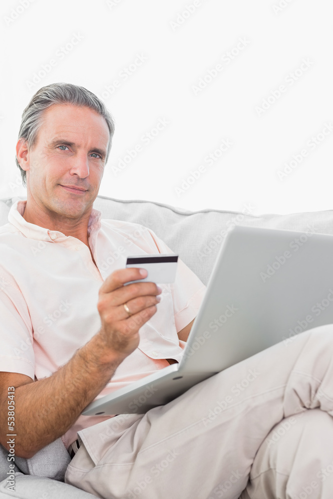 Man on his couch using laptop for shopping online smiling at cam Man on his couch using laptop for shopping online smiling at cam