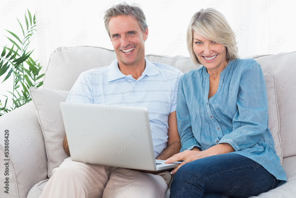 Happy couple using laptop together on the couch looking at camer Happy couple using laptop together on the couch looking at camer