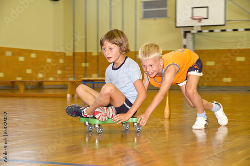 Photo shows a girl and a boy during a sport lesson in a gymnasium. The boy pushes his girlfriend on a roller board
