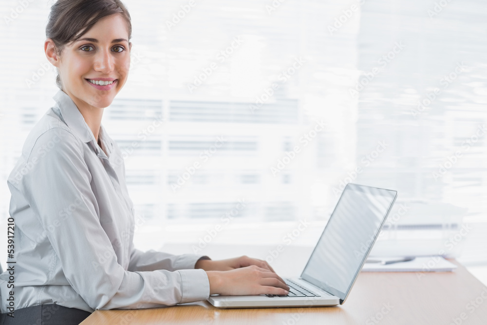Businesswoman typing on her laptop at desk and smiling at camera Businesswoman typing on her laptop at desk and smiling at camera