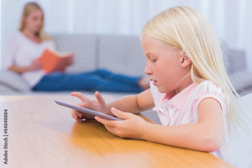 Little girl using tablet while her mother is reading on the couc Little girl using tablet while her mother is reading on the couc