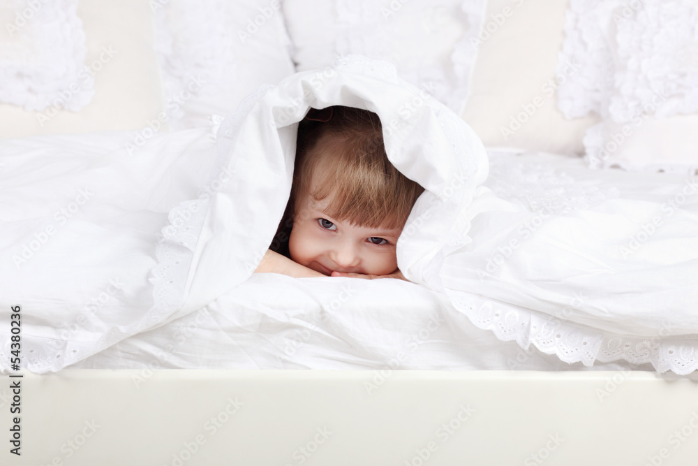 Little beautiful girl looks out from under warm blanket on bed. Little beautiful girl looks out from under warm blanket on bed.