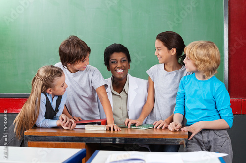 Teacher Sitting At Desk With Students At Desk