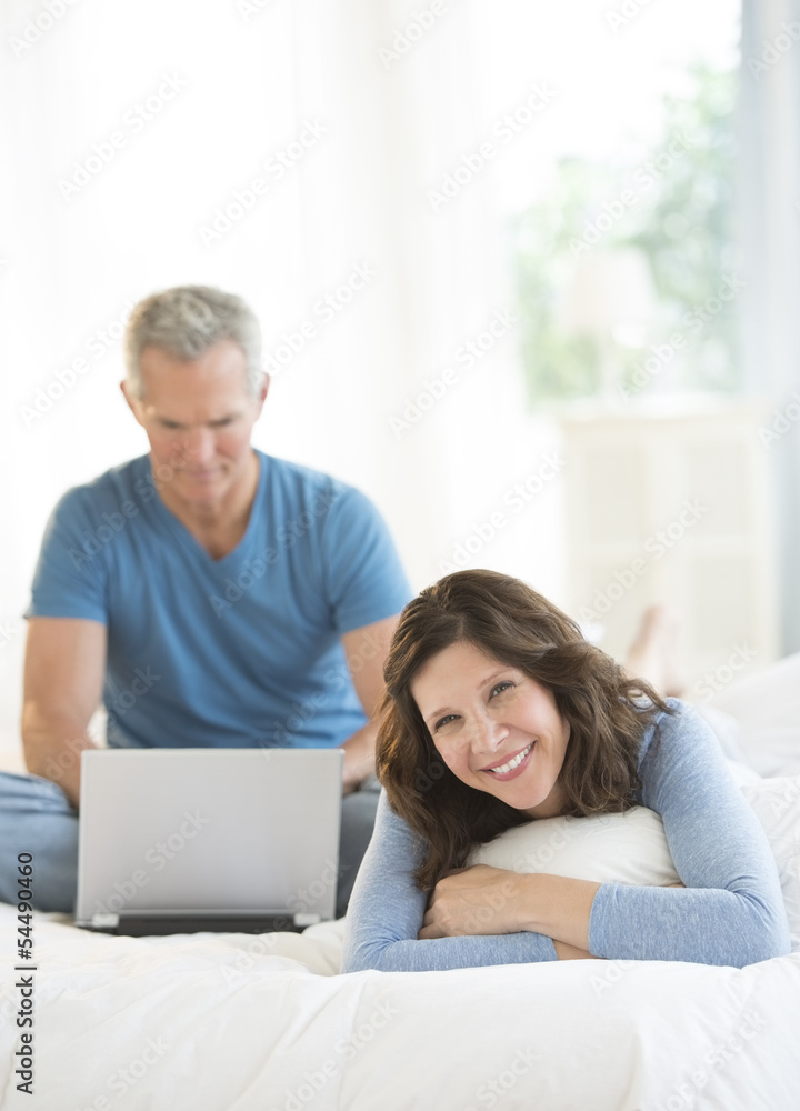Woman Relaxing In Bed While Man Using Laptop Woman Relaxing In Bed While Man Using Laptop
