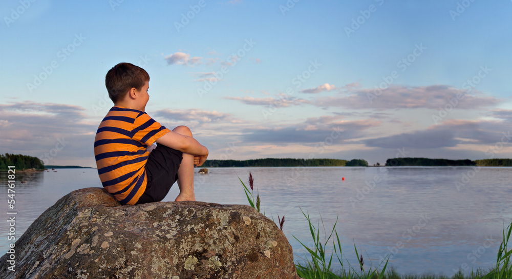 Obraz premium Panorama of a boy sitting on rock in summer evening sunlight
