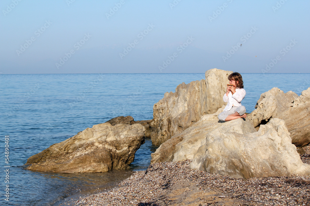 little girl sitting on a rock by the sea and playing pan pipe