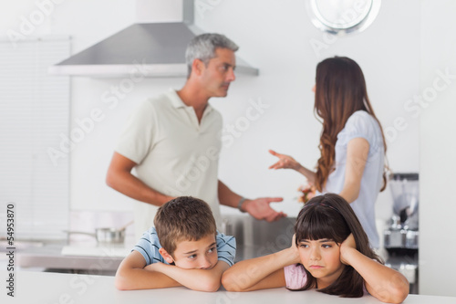 Unhappy siblings sitting in kitchen with their parents who are f
