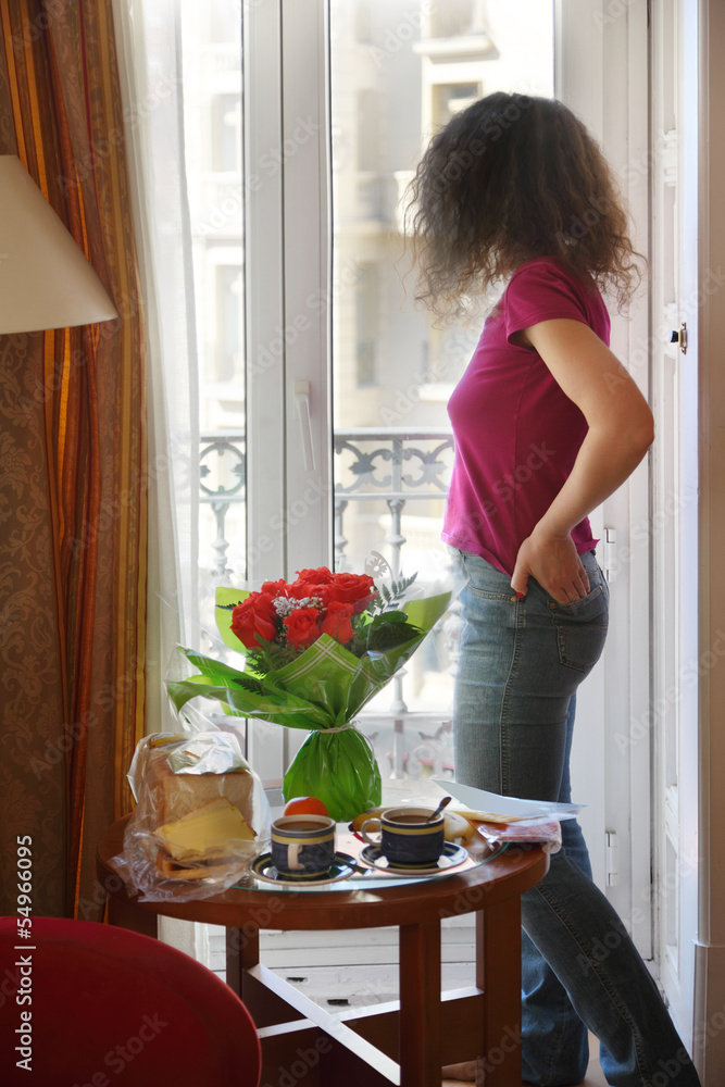 Young woman stands and looks out window at table Young woman stands and looks out window at table