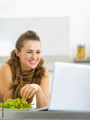 Smiling young woman eating grape and using laptop in kitchen