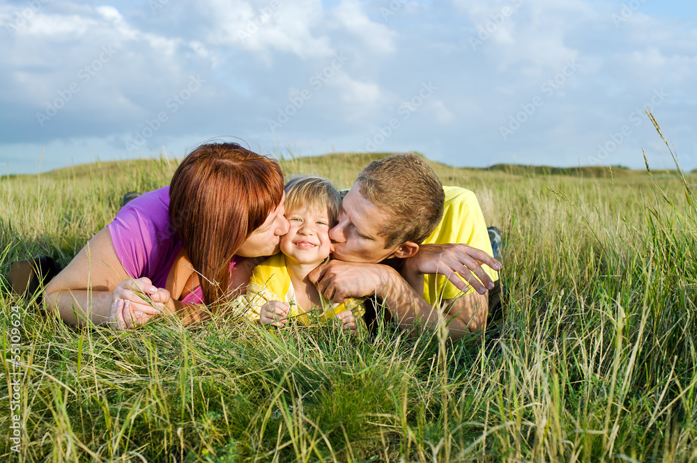 Fototapeta premium Parents kissing their daughter lying on a green meadow