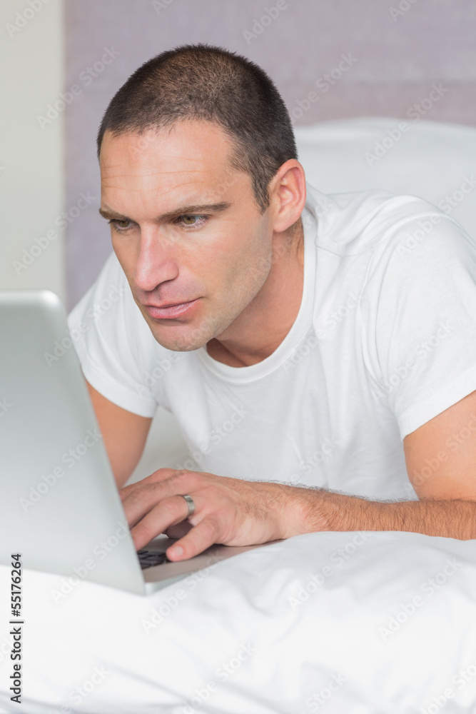Serious man lying on bed using his laptop Serious man lying on bed using his laptop