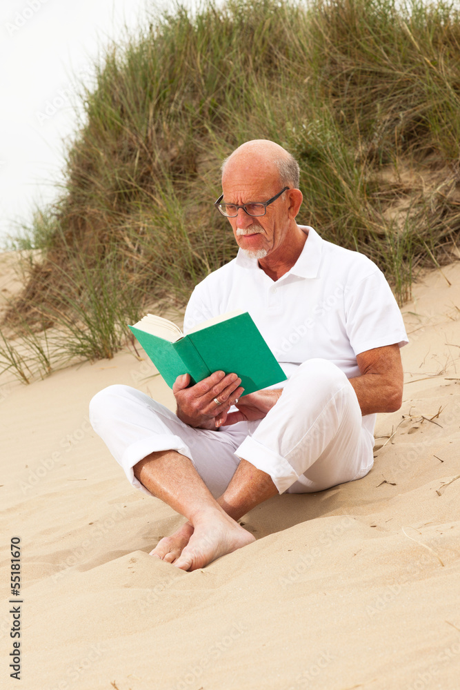 Retired man with beard and glasses reading a book in grass dune