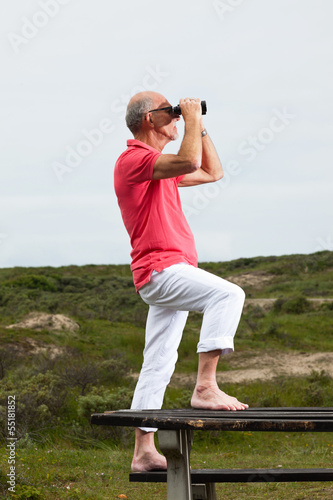 Retired man with beard and glasses using binoculars outdoors in