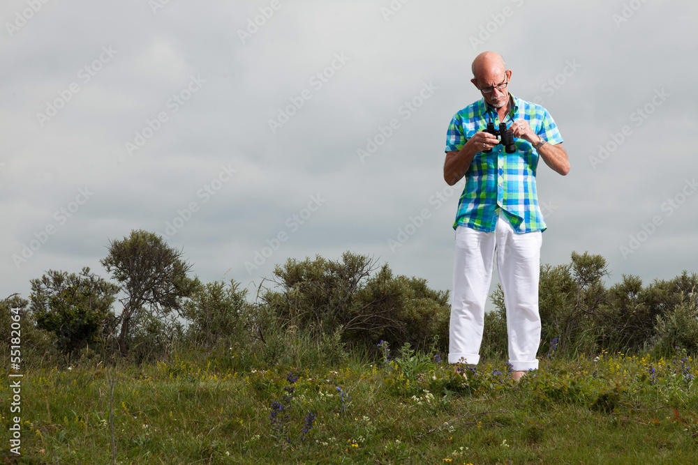 Senior man with beard and glasses using binoculars outdoors in g