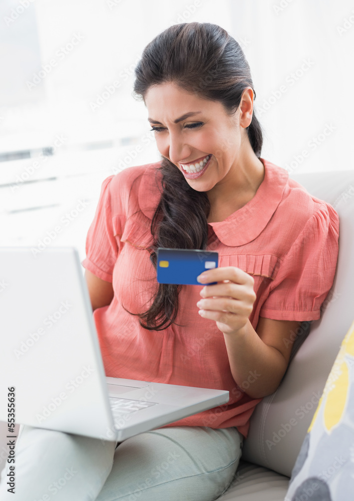 Cheerful brunette sitting on her sofa using laptop to shop onlin