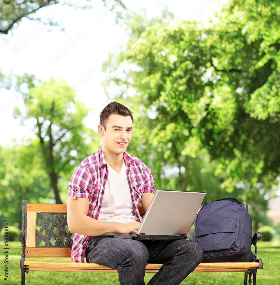 Smiling male student sitting on a bench and working on a laptop