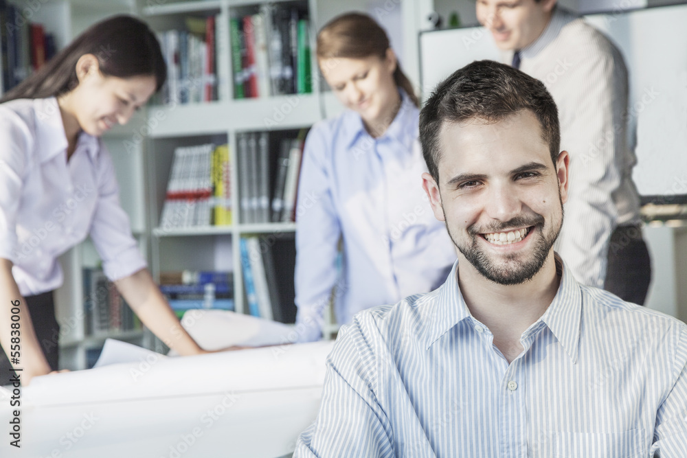 Smiling architect looking at camera while colleagues are planning around a table and looking at a blueprint Smiling architect looking at camera while colleagues are planning around a table and looking at a blueprint