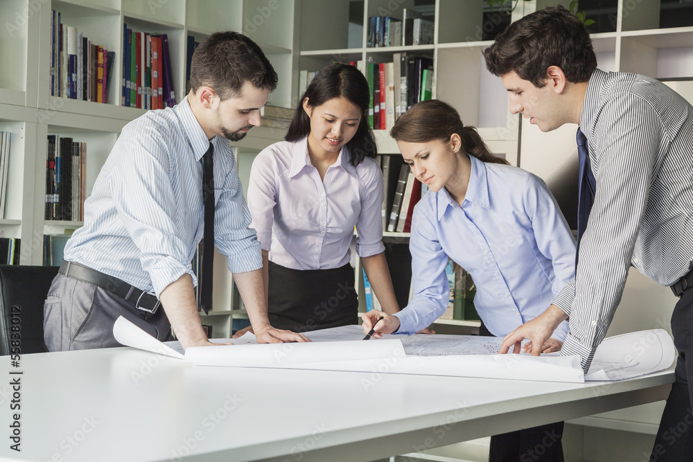 Four architects standing and planning around a table while looking down at blueprint Four architects standing and planning around a table while looking down at blueprint