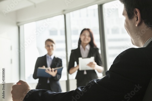 Two businesswomen standing up and presenting during a business meeting as a businessman watches