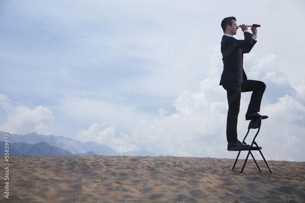 Businessman standing on a chair and looking through a telescope in the middle of the desert 