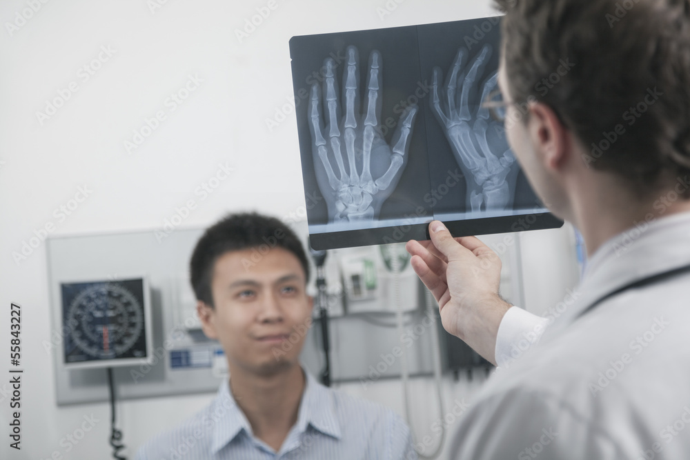 Doctor holding up and looking at x-ray of patients hand bones, patient in the background Doctor holding up and looking at x-ray of patients hand bones, patient in the background