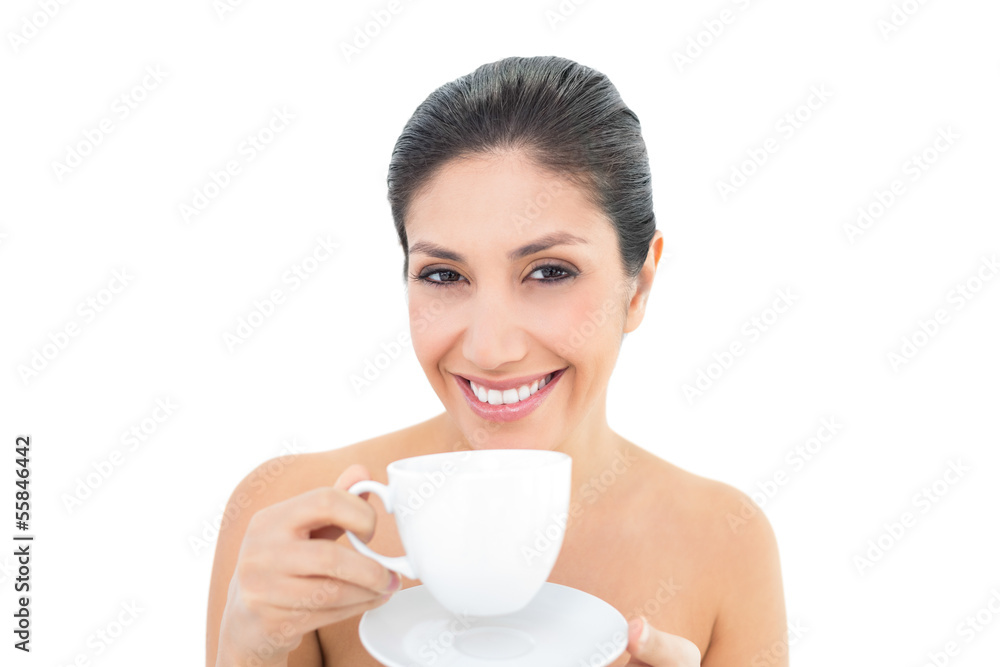 Attractive brunette holding a cup and saucer and smiling at came Attractive brunette holding a cup and saucer and smiling at came