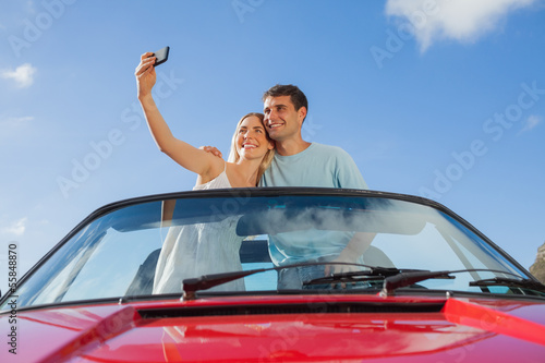 Cheerful couple standing in red cabriolet taking picture