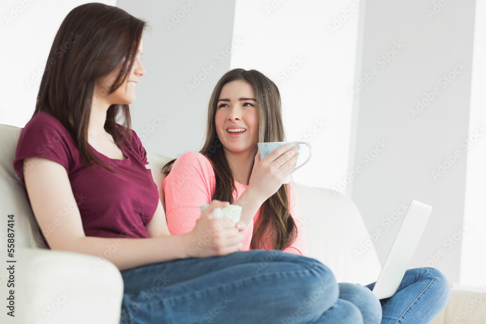 Two laughing girls sitting on a sofa enjoying a beverage and hol Two laughing girls sitting on a sofa enjoying a beverage and hol