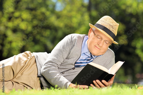 Senior man lying on a grass and reading a book in a park