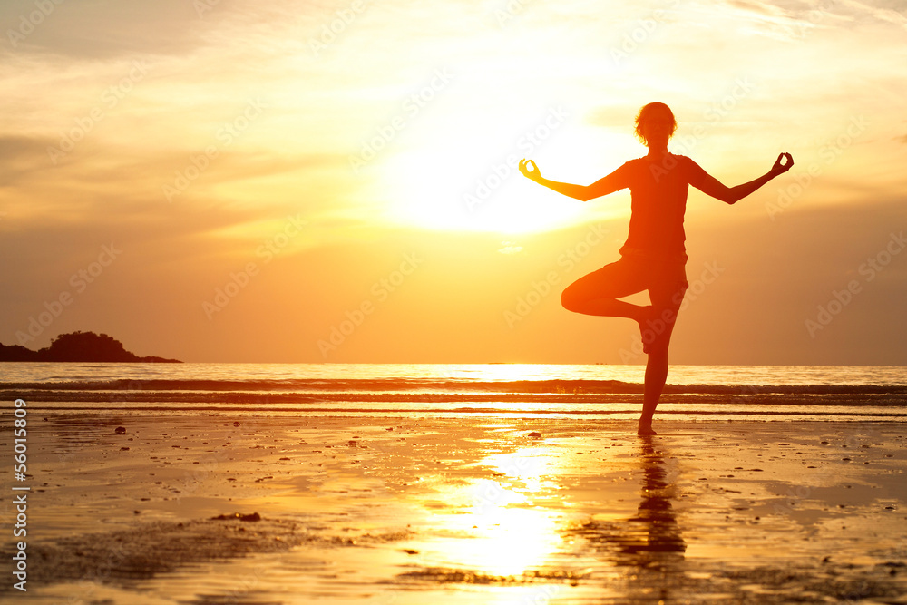 Backlit, young woman practicing yoga on the beach at sunset.