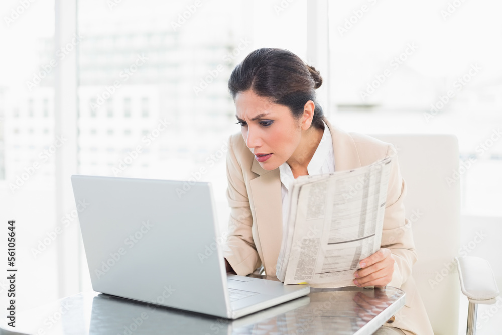 Stern businesswoman holding newspaper while working on laptop Stern businesswoman holding newspaper while working on laptop