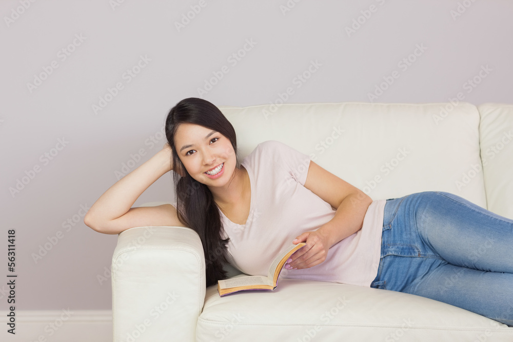 Happy asian girl lying on the sofa reading book smiling at camer Happy asian girl lying on the sofa reading book smiling at camer