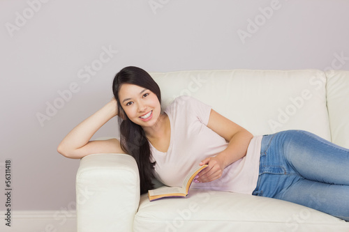 Happy asian girl lying on the sofa reading book smiling at camer