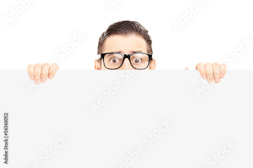 Scared young man with glasses hiding behind a blank panel