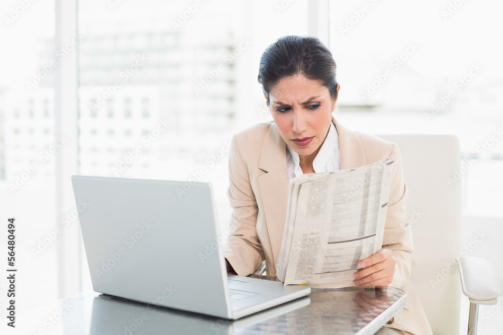Serious businesswoman reading newspaper while working on laptop Serious businesswoman reading newspaper while working on laptop