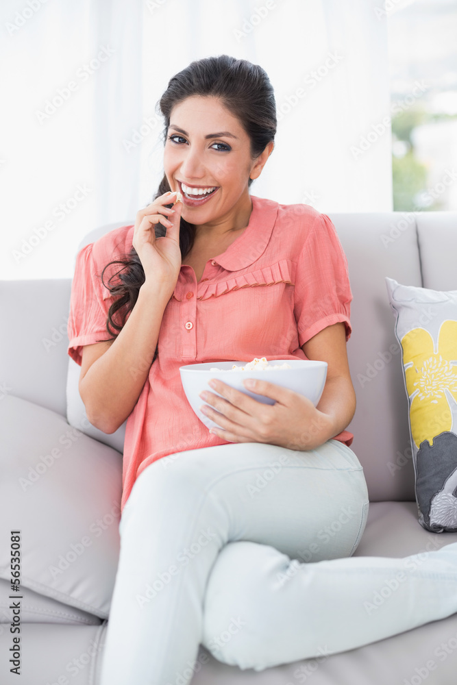 Cheerful brunette sitting on her sofa watching tv eating bowl of Cheerful brunette sitting on her sofa watching tv eating bowl of