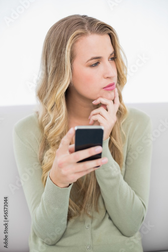 Pensive pretty blonde sending a text sitting on cosy sofa