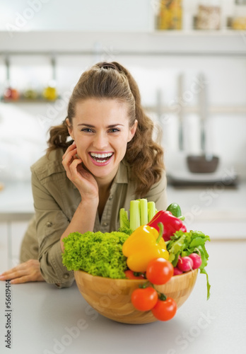 Happy young housewife near plate full of vegetables in kitchen