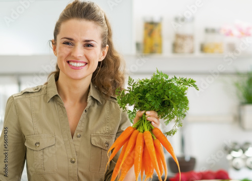 Happy young housewife holding bunch of carrots in kitchen