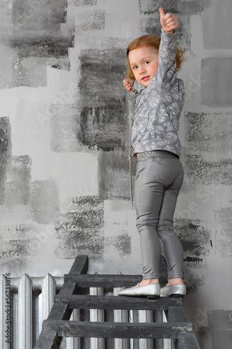 A little girl with hand raised standing on ladder at gray wall photo