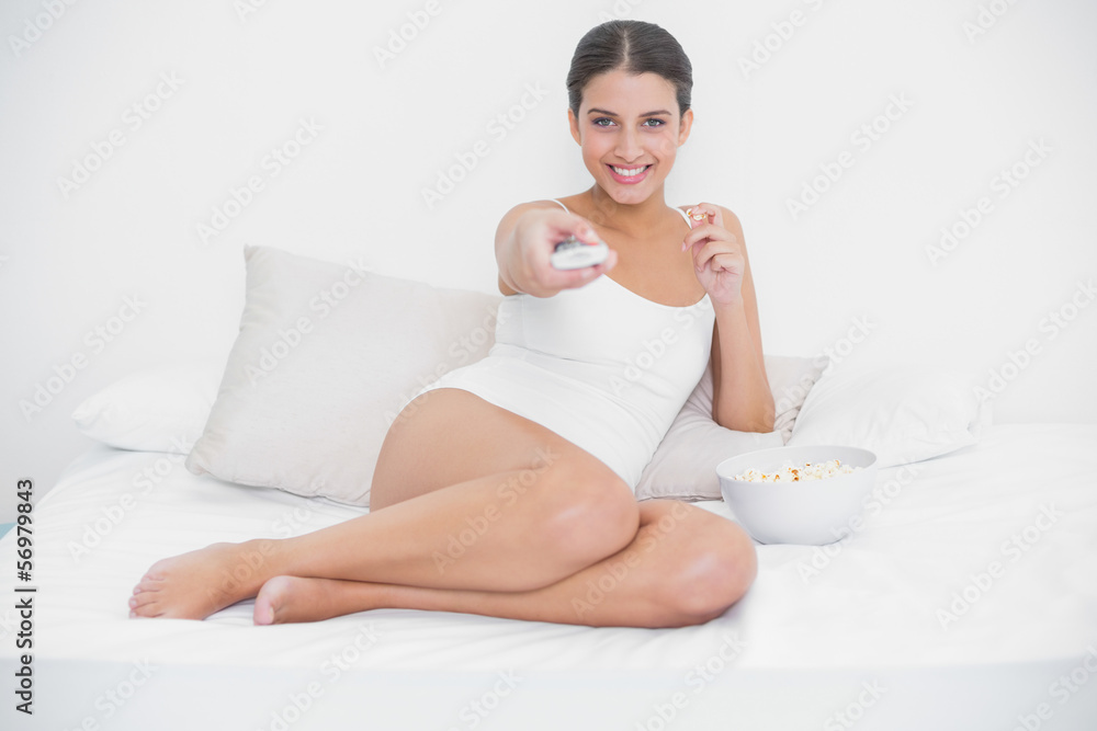 Cheerful young brown haired model in white pajamas watching tv w Cheerful young brown haired model in white pajamas watching tv w