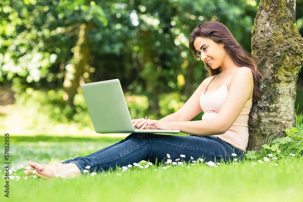 Cute smiling woman leaning against a tree in a park using her la