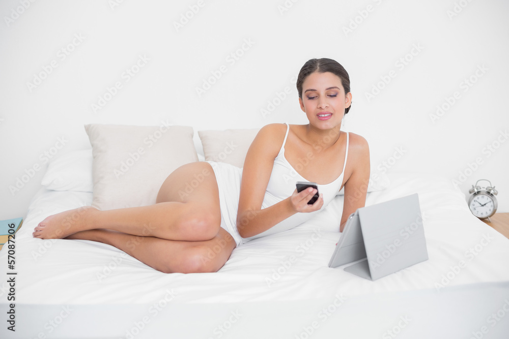 Thinking young brown haired model in white pajamas using a mobil Thinking young brown haired model in white pajamas using a mobil