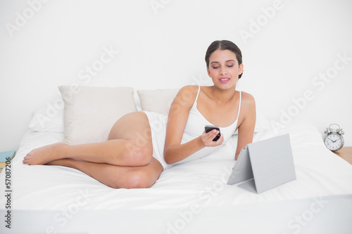 Thinking young brown haired model in white pajamas using a mobil
