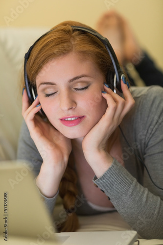 Calm redhead lying on the sofa with her laptop listening to musi