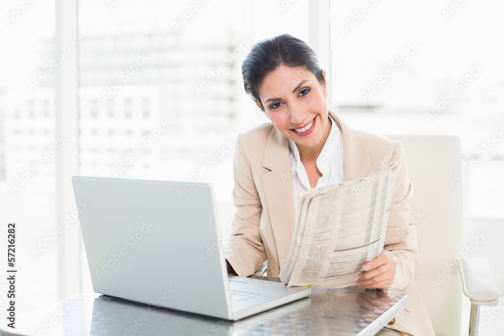 Smiling businesswoman holding newspaper while working on laptop Smiling businesswoman holding newspaper while working on laptop