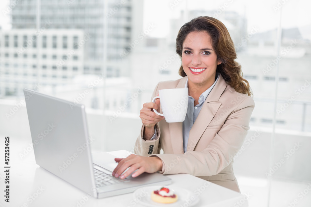 Smiling businesswoman using laptop at her desk and holding mug Smiling businesswoman using laptop at her desk and holding mug
